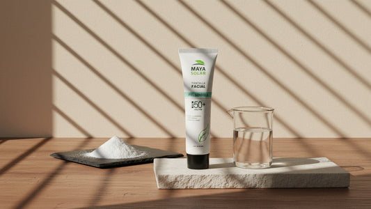 Pantalla Facial Piel Sensible FPS 50+ de Maya Solar entre un plato oscuro con polvo mineral blanco y un vaso de vidrio con líquido transparente, sobre una losa de piedra caliza