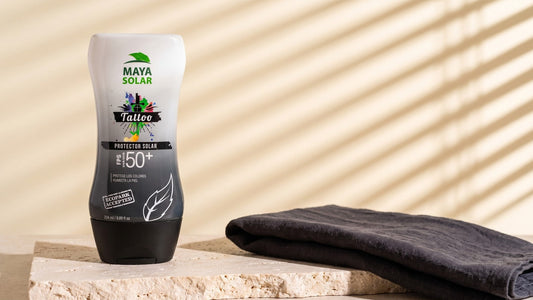 Bloqueador Solar para Tattoos FPS 50+ de Maya Solar de 204 ml de pie sobre una losa de piedra caliza, junto a una tela gris oscura doblada, con la luz de una persiana rayando la pared